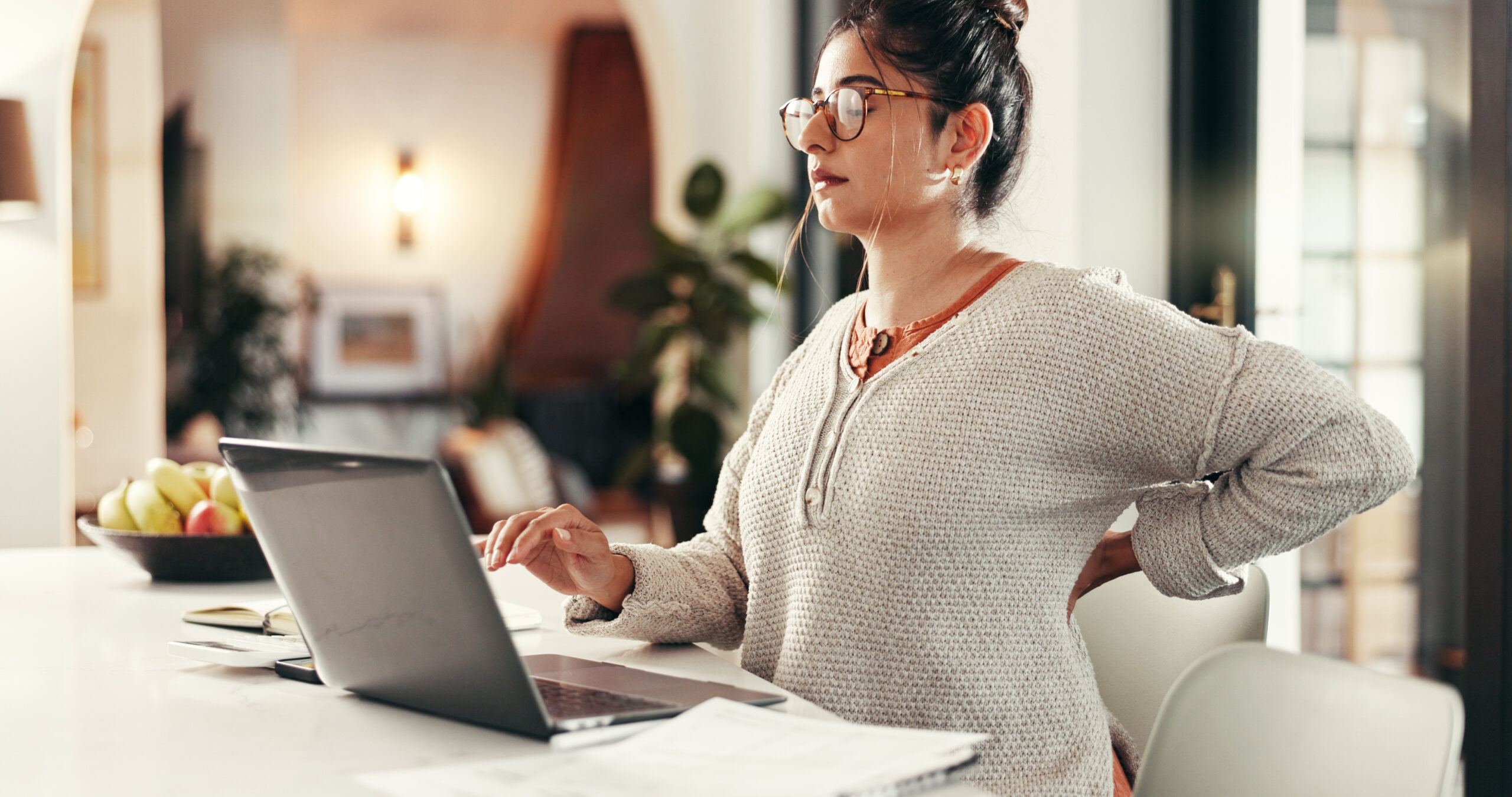 Woman sitting at a laptop with back pain, holding her lower back due to poor posture.